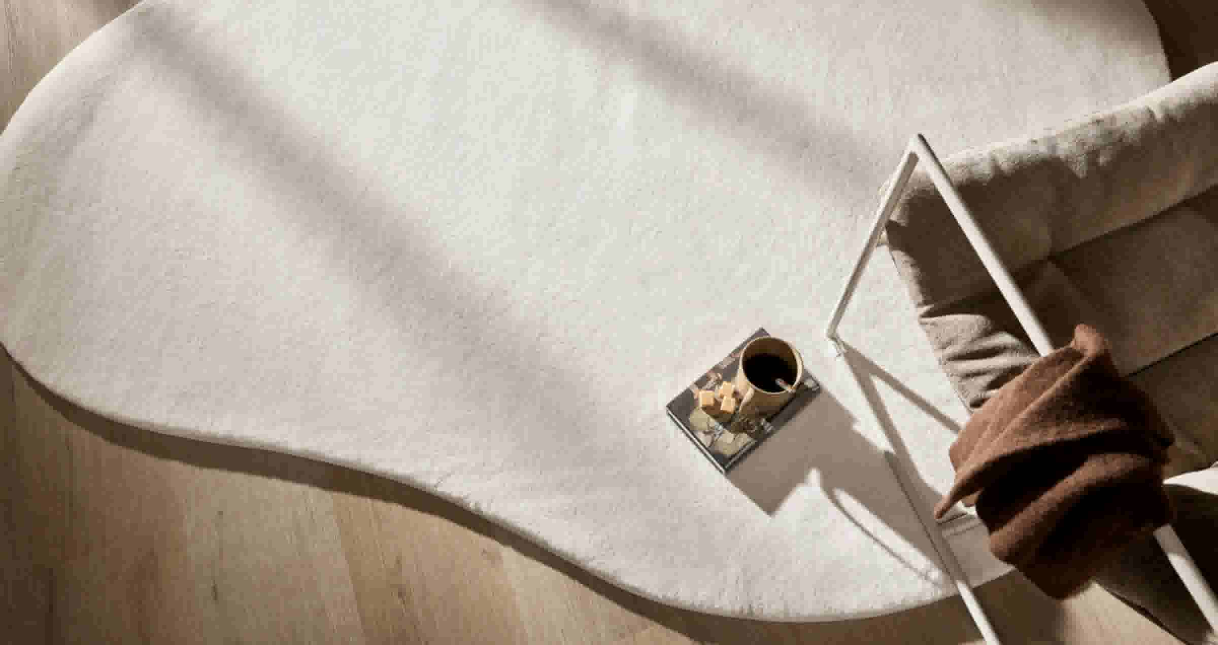 Minimalistisch interieur met wit tapijt, witte stoel met bruine deken en een bijzettafel met koffiekopje en snacks.