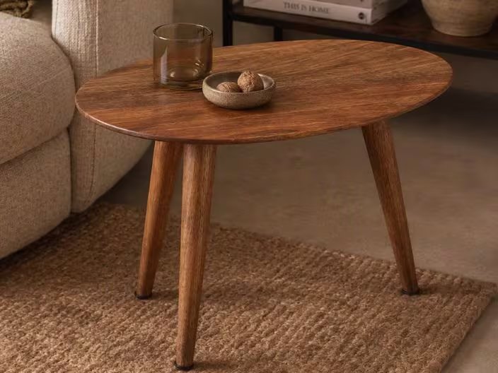 Ovale houten salontafel met drie poten op een juten vloerkleed, met een glas en schaaltje noten erop, naast een beige bank.