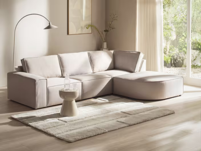 Moderne beige modulaire hoekbank James in lichte woonkamer met staande lamp, bijzettafel en grote ramen die uitkijken op een groene tuin.