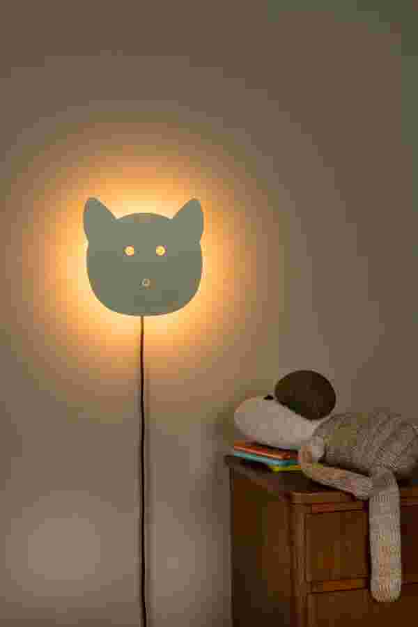 Wandlamp van Lucide in kattenvorm met warme gloed op de muur, naast een houten kastje met knuffels en boekjes.