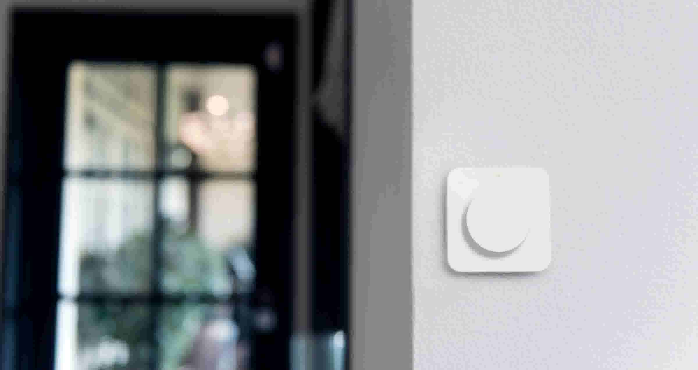 Witte slimme schakelaar met ronde knop op een grijze muur, met op de achtergrond een wazige donkere deur en raam.