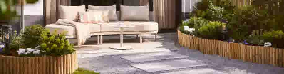 Stijlvolle tuin met beige loungeset, salontafel, stapstenen, grind en houten borders met bloemen en planten.