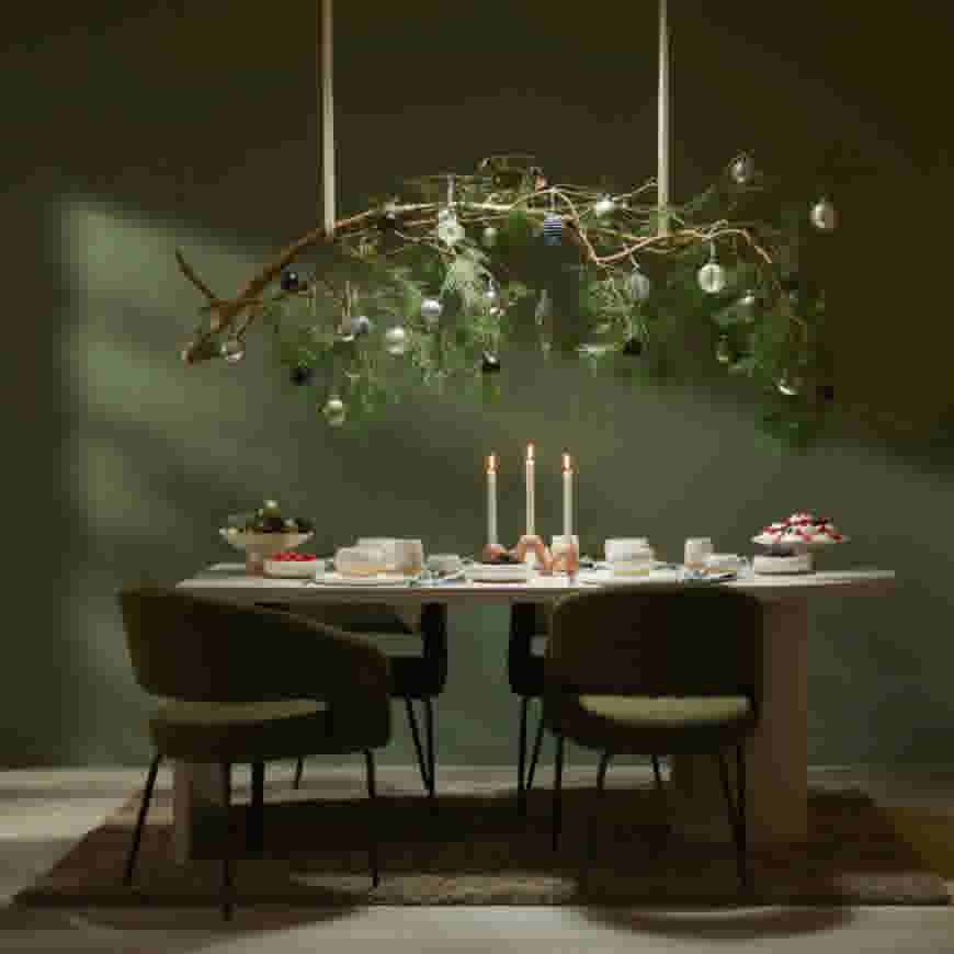 Feestelijke eettafel met kaarsen en gebak onder een hangende dennentak versierd met kerstballen tegen een groene muur.