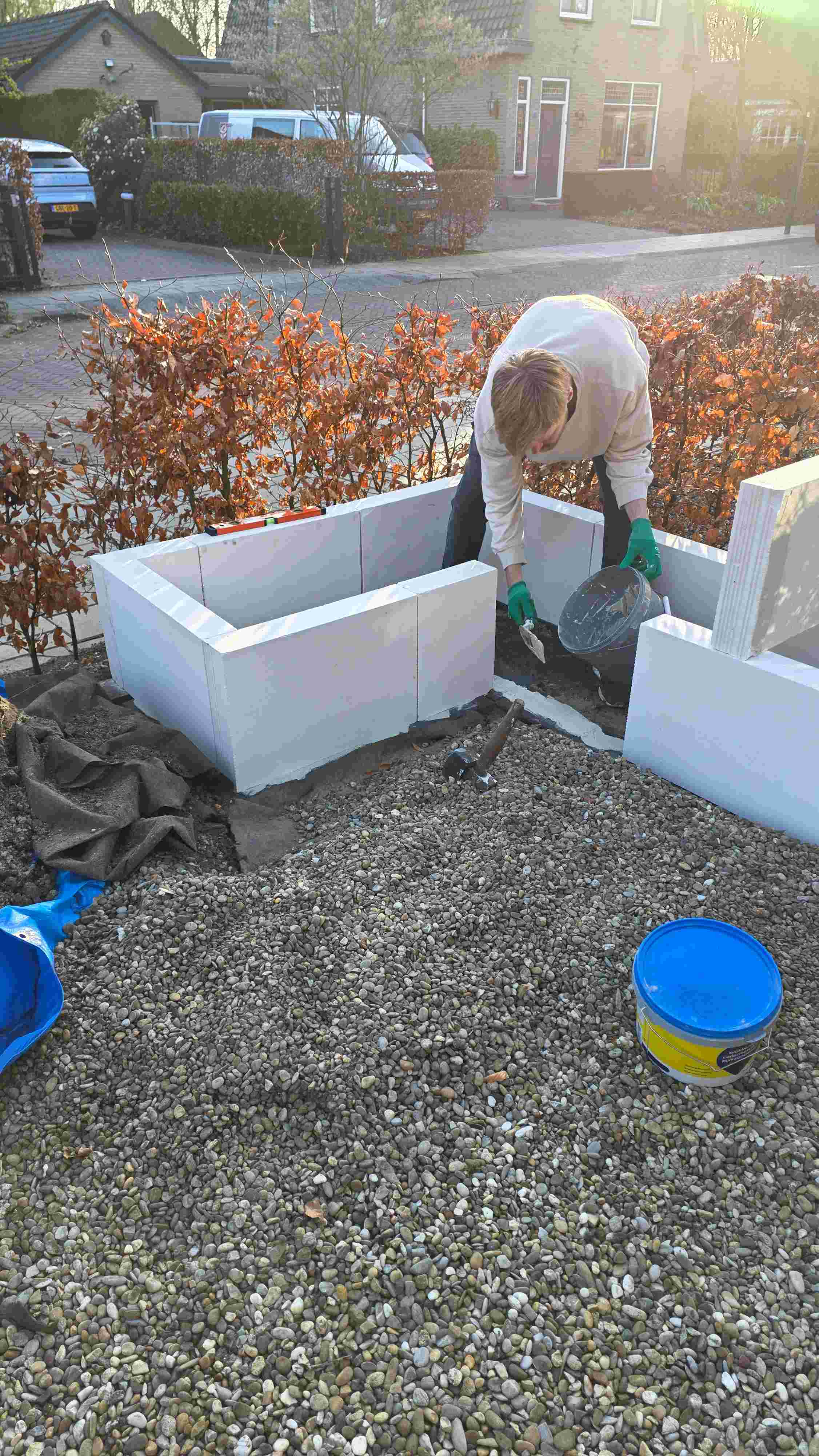 Persoon met groene handschoenen bouwt witte bakken in een tuin met grind, naast oranje hagen en met huizen op de achtergrond.