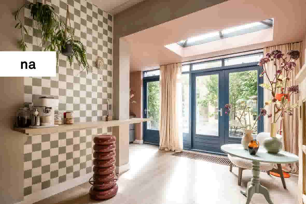 Moderne entree met geblokte muur, hangende planten, glazen deuren naar tuin, en mintgroen tafeltje met bloemen.