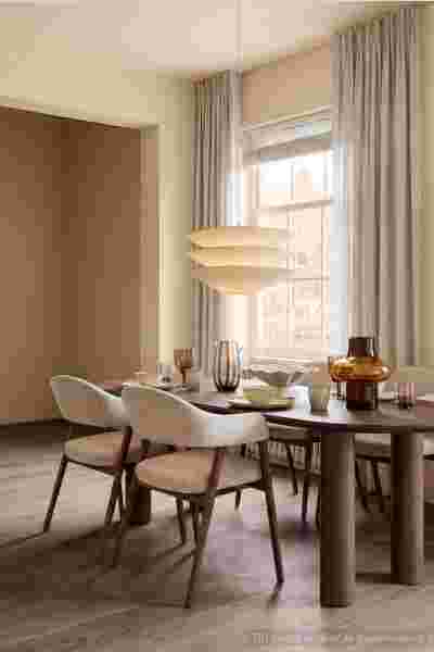 Moderne eetkamer met beige stoelen, houten tafel, hanglamp en grijze gordijnen bij groot raam met natuurlijk licht.