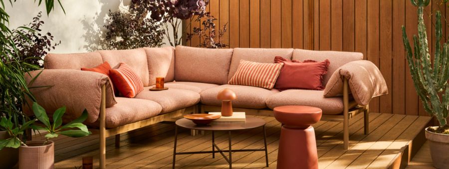 Gezellige roze loungebank met terracotta kussens op houten terras, omringd door planten en decoratieve bijzettafels.