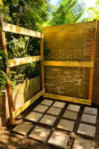 Houten frame tegen een bakstenen muur in een tuin, met betonnen vlonders op de grond en groene bomen op de achtergrond.