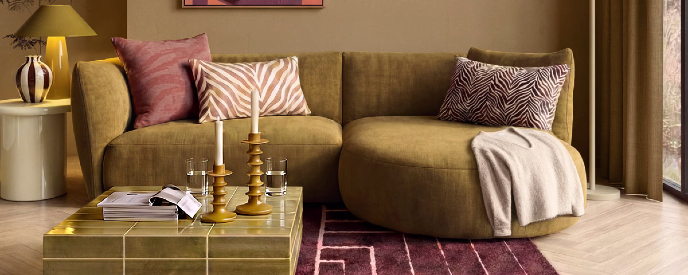 Moderne woonkamer met olijfgroene bank, roze en zebraprint kussens, gouden salontafel en abstract kunstwerk aan beige muur.