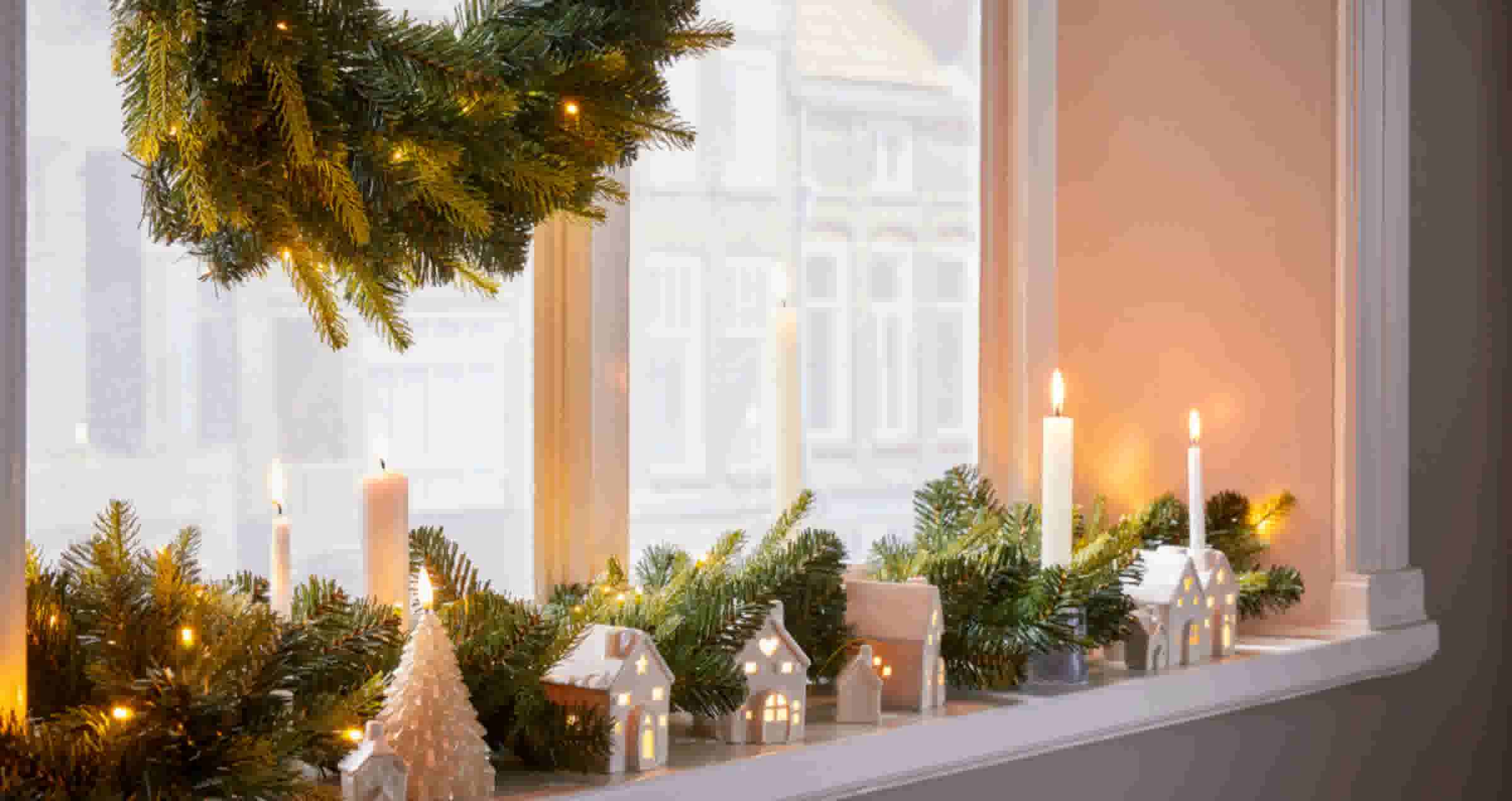Een vensterbank versierd met een kerstkrans, dennegroen, brandende kaarsen en kleine witte huisjes met lichtjes erin.