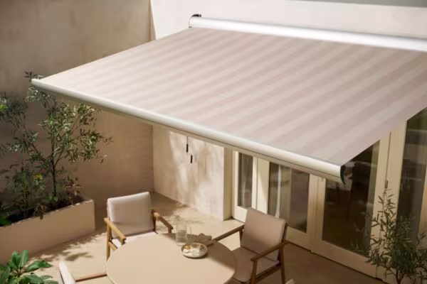 Uitschuifbaar zonnescherm boven terras met tafel en stoelen, omringd door planten tegen een lichte muur.