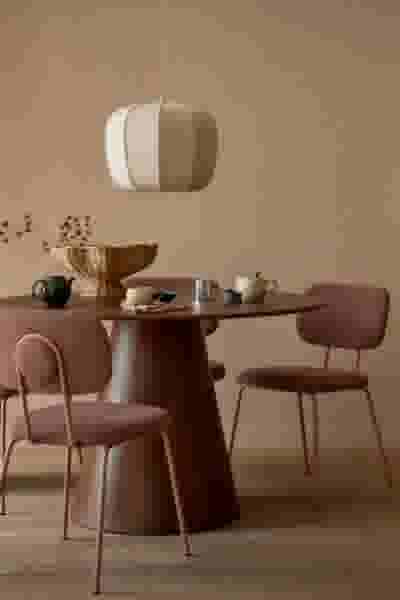 Ronde bruine eettafel met roze stoelen, theegerei en een rieten mand. Crèmekleurige hanglamp erboven. Warme, neutrale tinten.