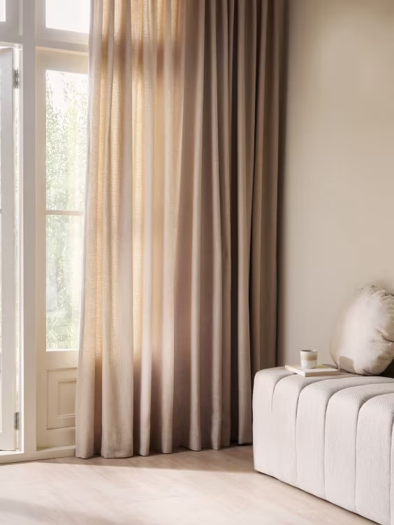 Lichte kamer met beige en taupe gordijnen voor hoge ramen, witte bank en neutrale wandkleuren.