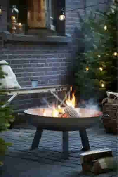 Gezellige vuurschaal met brandend hout op terras met kerstboom, bakstenen muur en sfeervolle lichtslingers.