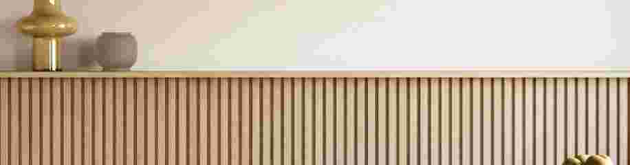 Houten dressoir met verticale latten, waarop een gouden lamp en beige vaas staan, tegen een lichte muur.