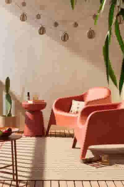 Gezellige tuin met koraalkleurige kuipstoelen, decoratieve lichtslingers, groene planten en een tapijt in warme tinten.