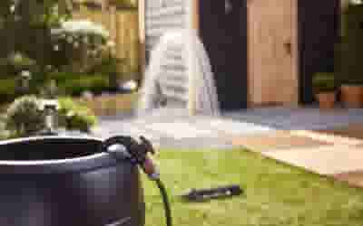 Zwarte tuinslang aangesloten op een pot, met een sproeier die water verspreidt in een achtertuin met gras en planten.