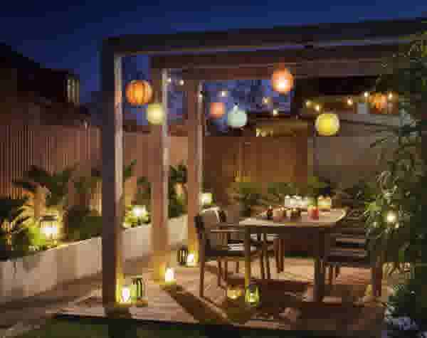 Sfeervolle tuin in de avond met pergola, kleurrijke lampionnen, lichtjes en een eettafel omringd door planten en lantaarns.