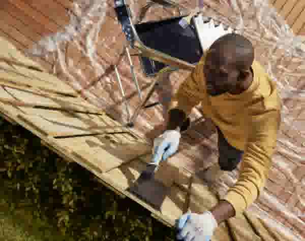 Man in geel sweatshirt schildert houten planken met een kwast, draagt witte handschoenen, gezien van bovenaf buiten.