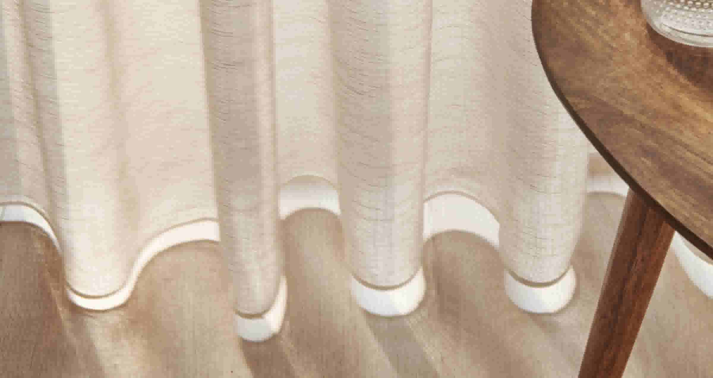 Crèmekleurig gordijn dat net boven een houten vloer valt, met rechts een glimp van een houten tafel of meubel.