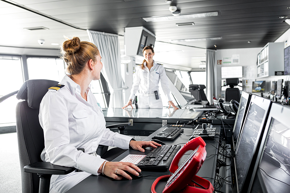 Schiffsbrücke. Zwei weibliche Offizierinnen arbeiten auf der Brücke und lächeln sich dabei an. 