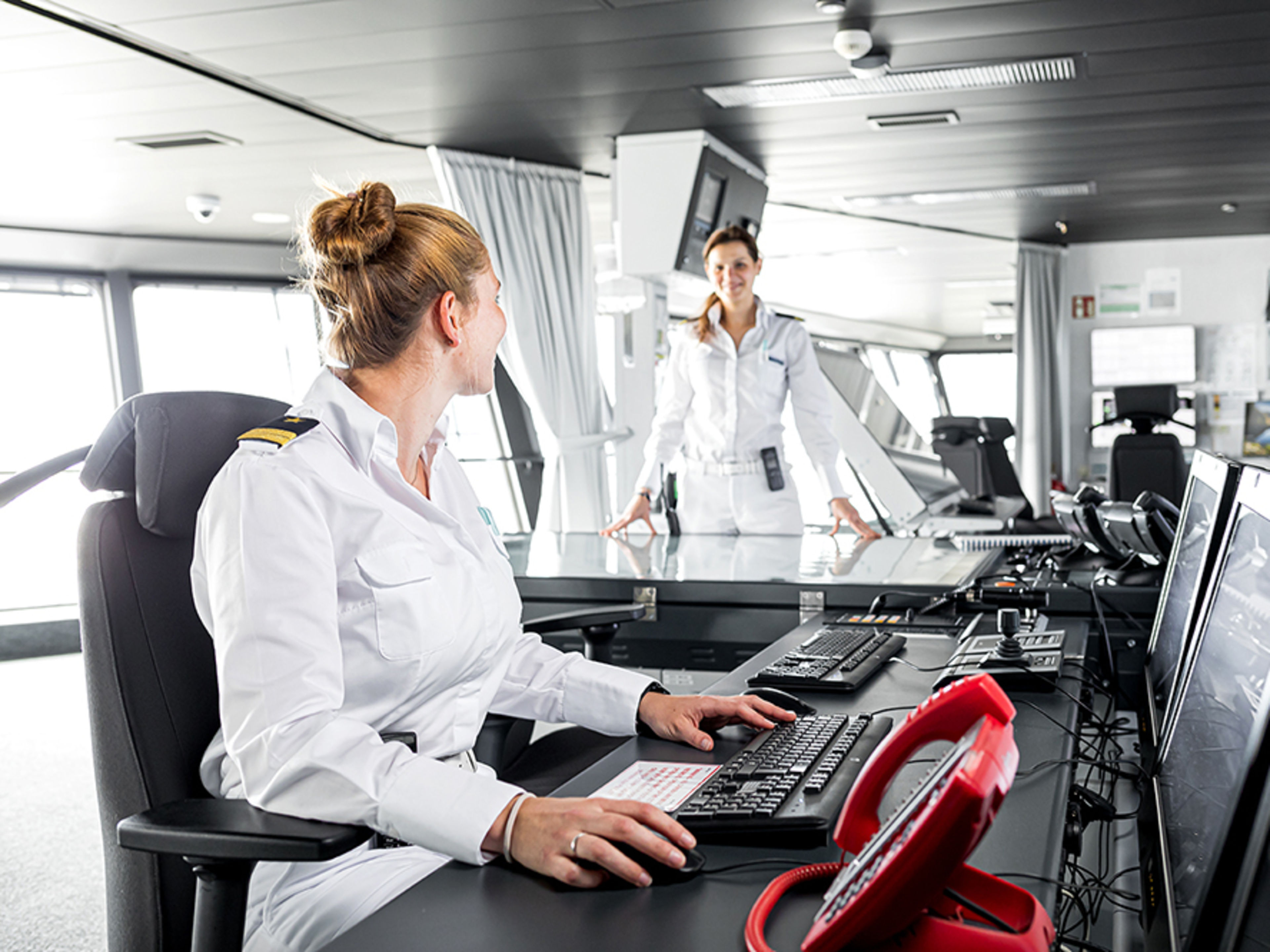 Schiffsbrücke. Zwei weibliche Offizierinnen arbeiten auf der Brücke und lächeln sich dabei an.