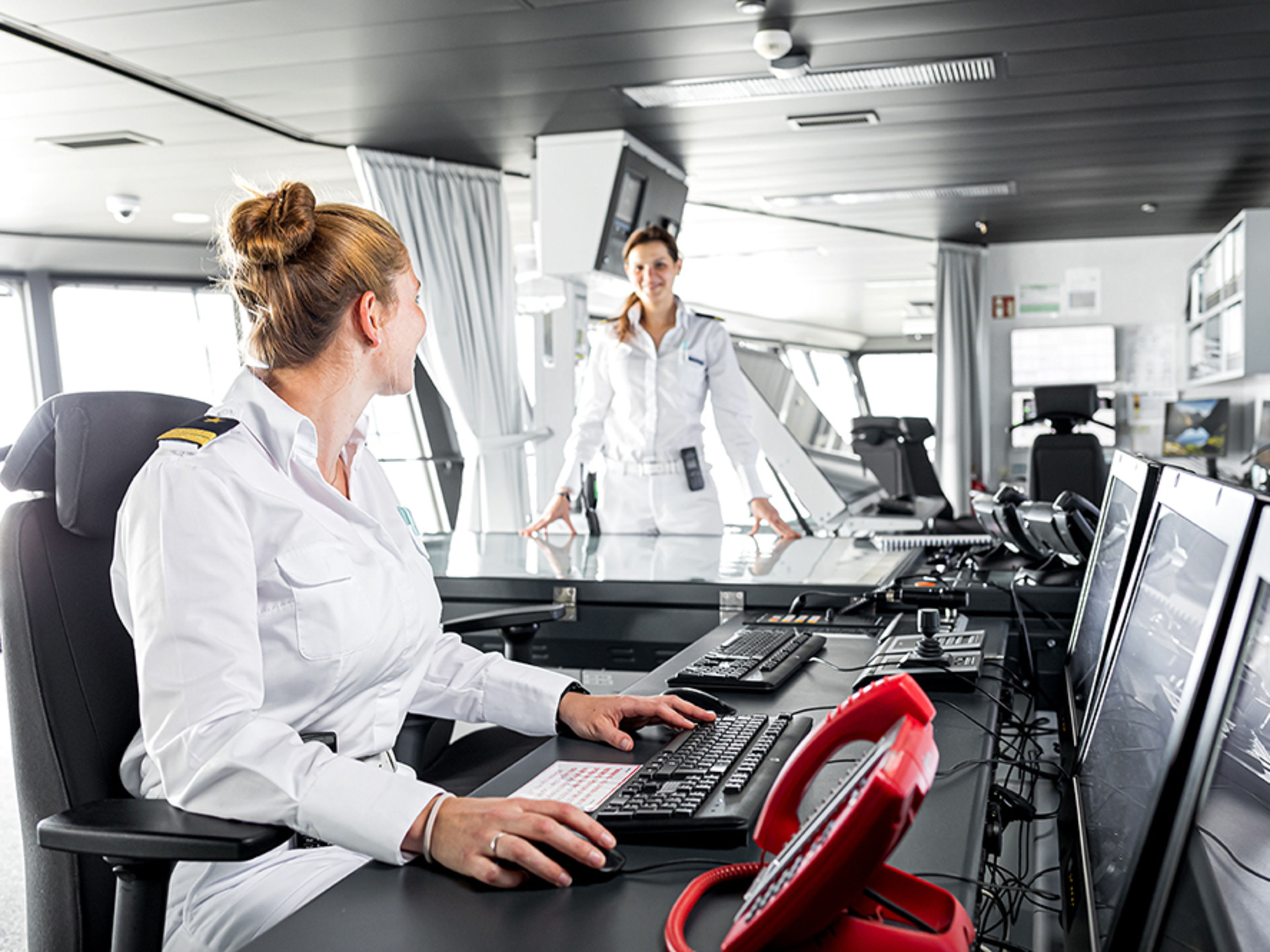 Schiffsbrücke. Zwei weibliche Offizierinnen arbeiten auf der Brücke und lächeln sich dabei an.