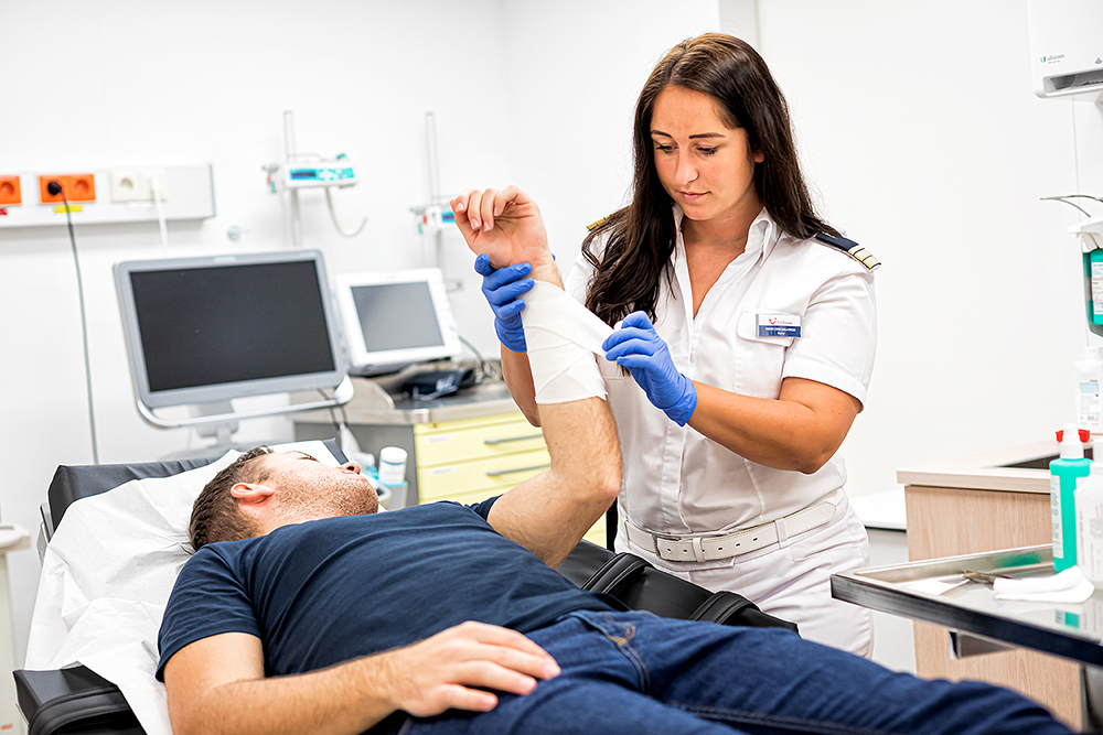Im Behandlungszimmer. Die weibliche Nurse verbindet das Handgelenk eines männlichen Patienten, der auf einer Liege liegt. 