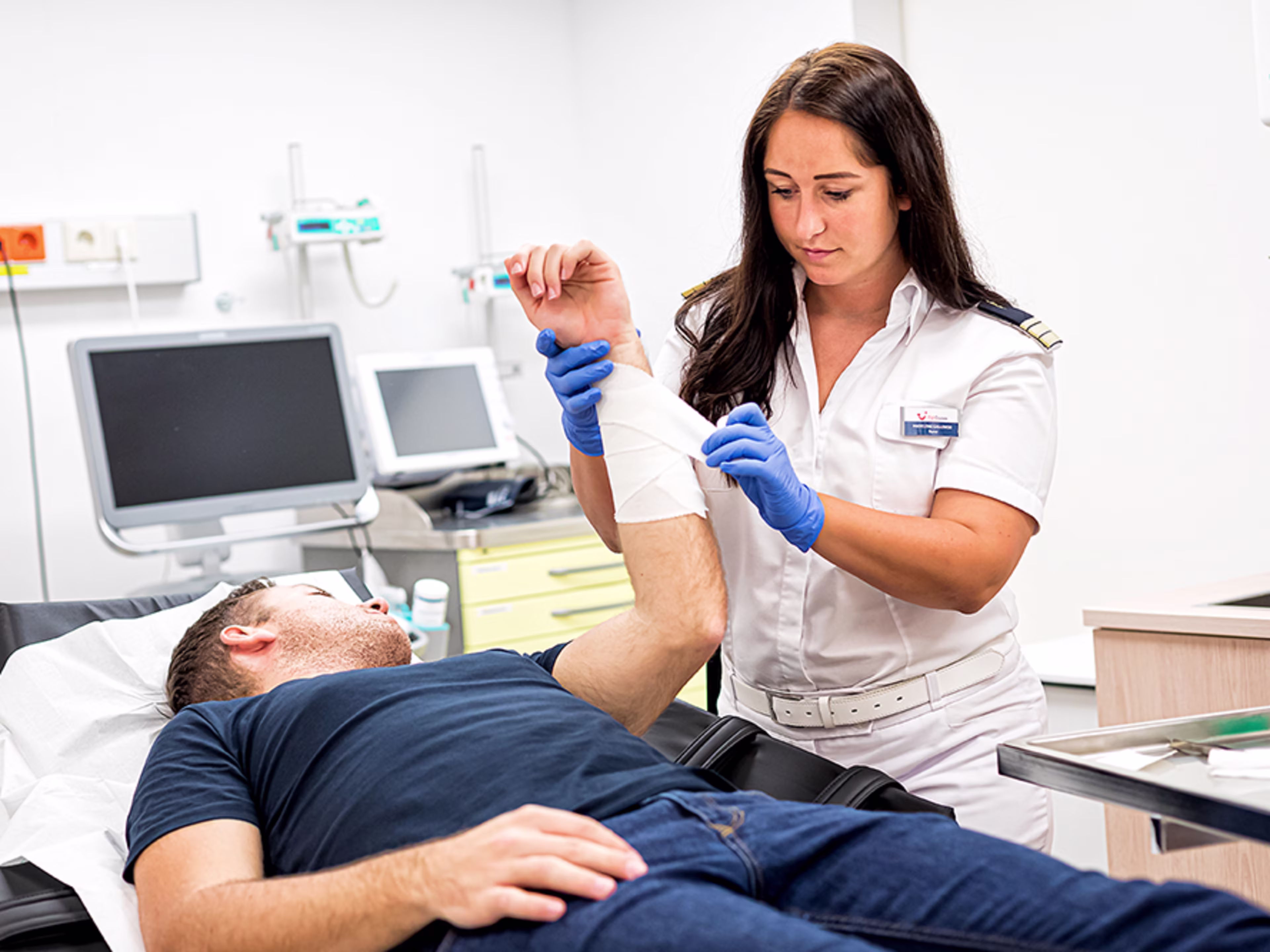 Im Behandlungszimmer. Die weibliche Nurse verbindet das Handgelenk eines männlichen Patienten, der auf einer Liege liegt.