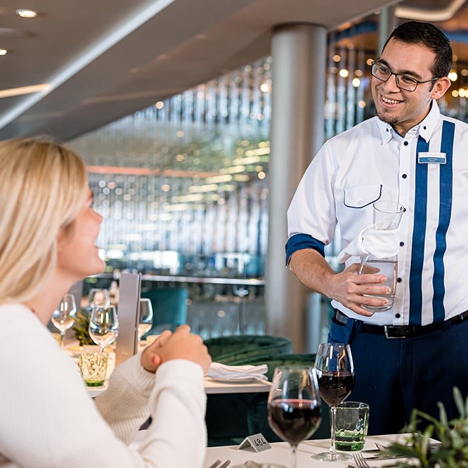 Restaurantbesuch. Ein freundlicher Kellner schenkt bei einem weiblichen Gast Wasser ein. Die beiden Personen lächeln sich an.