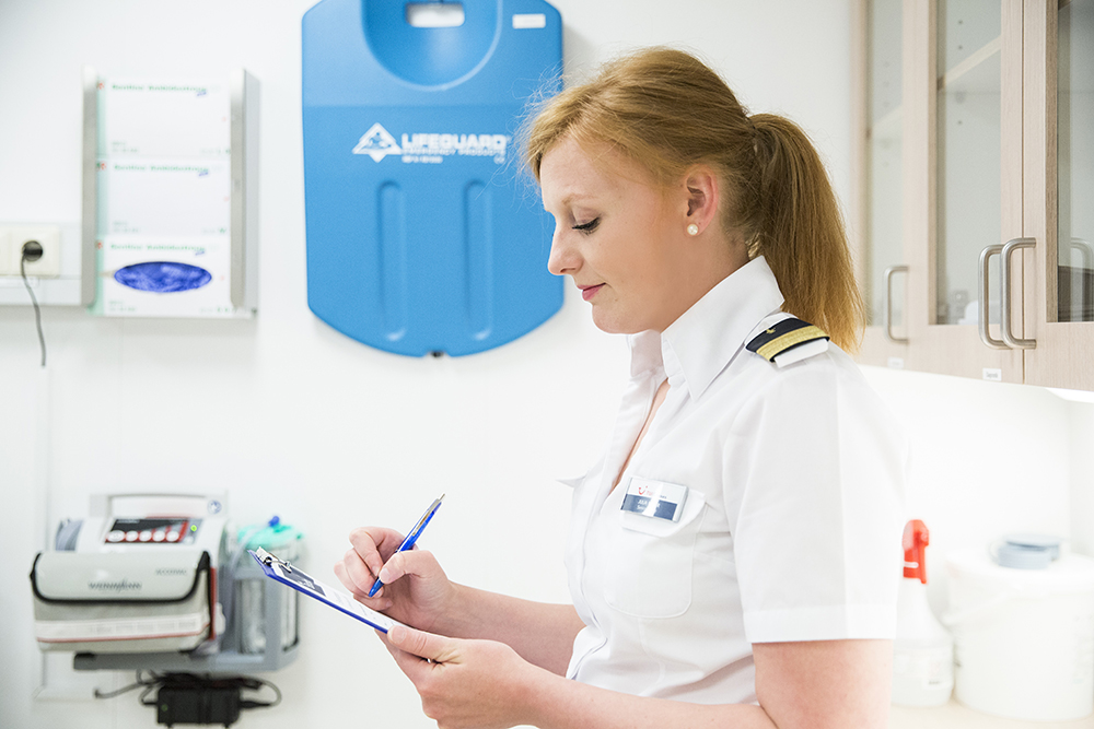 Im Bordhospital. Eine weibliche Crew Member wird von der Seite porträtiert, sie hat ein Klemmbrett in der Hand, auf welchem sie etwas ausfüllt. 