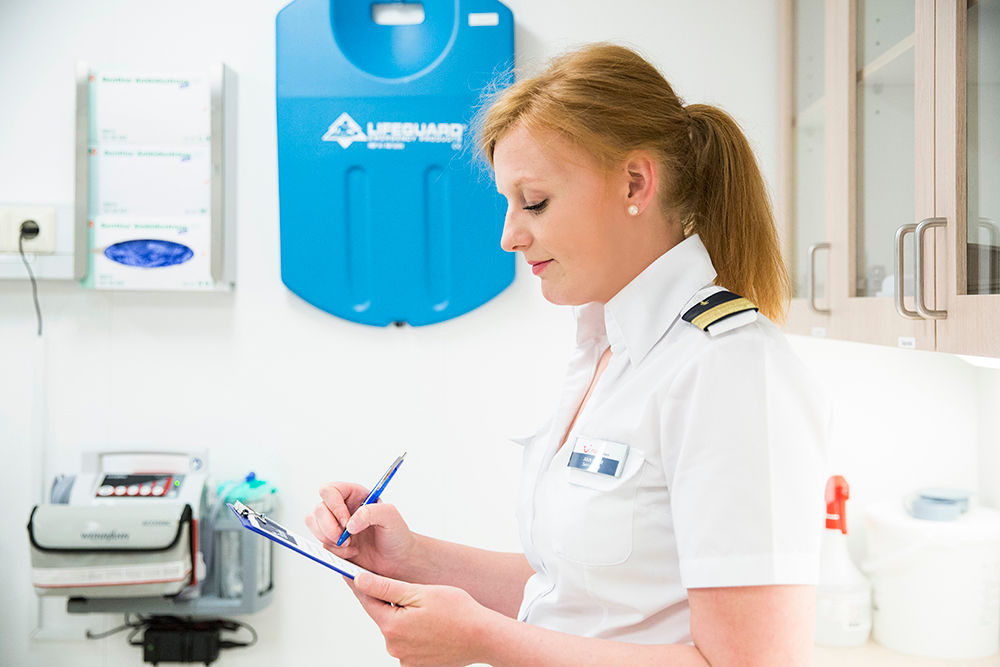 Im Bordhospital. Eine weibliche Crew Member wird von der Seite porträtiert, sie hat ein Klemmbrett in der Hand, auf welchem sie etwas ausfüllt. 