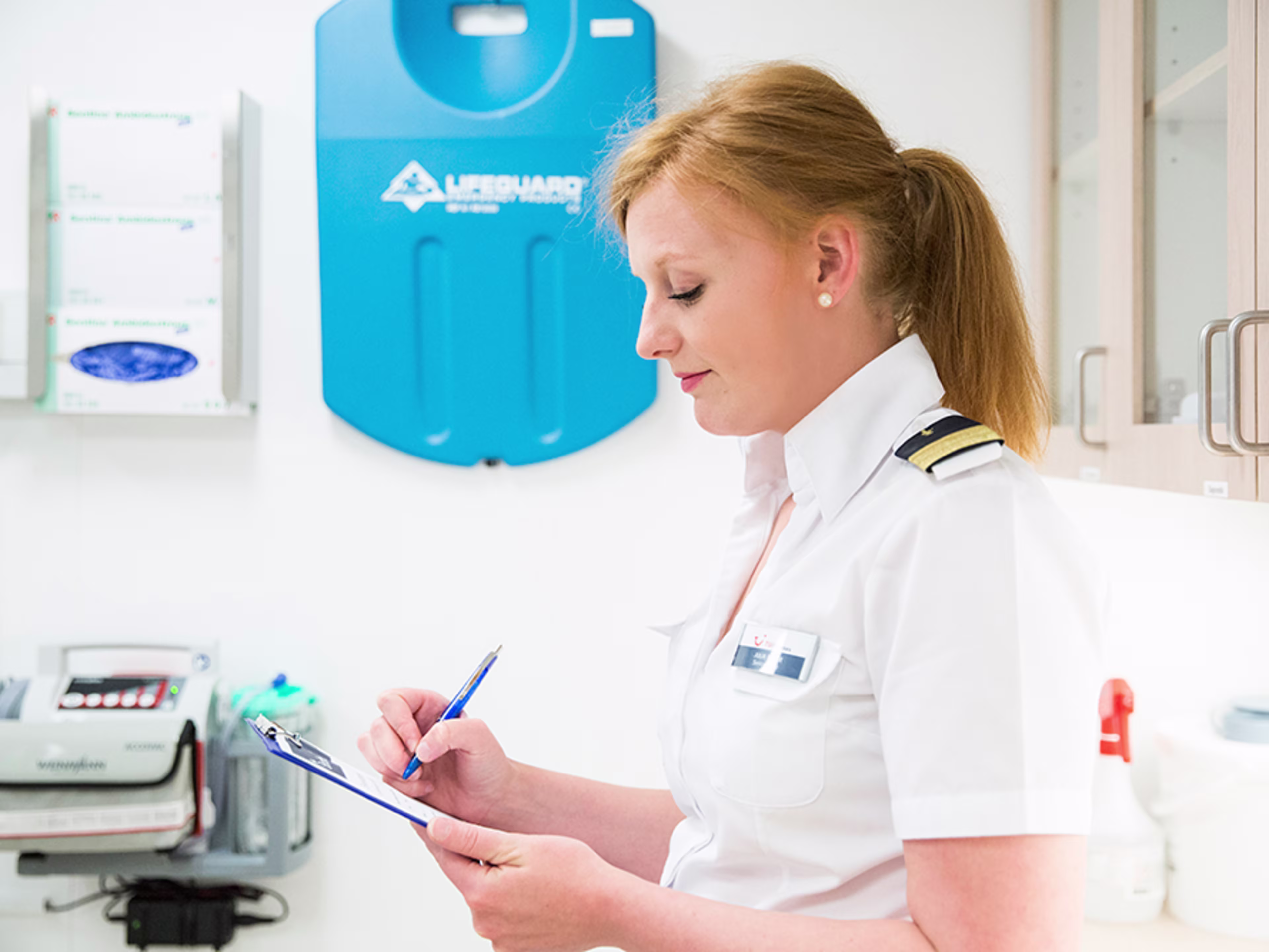 Im Bordhospital. Eine weibliche Crew Member wird von der Seite porträtiert, sie hat ein Klemmbrett in der Hand, auf welchem sie etwas ausfüllt.