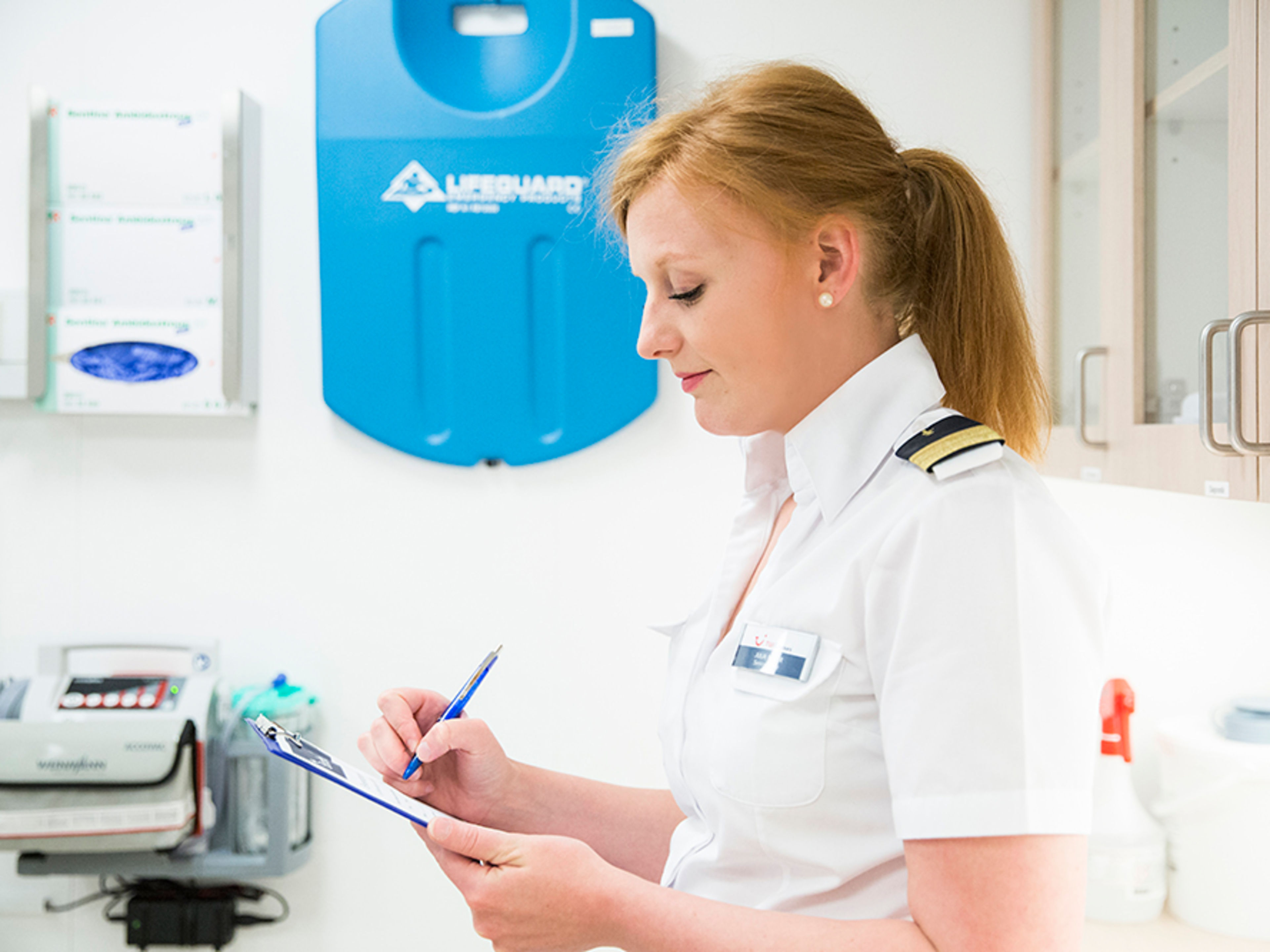Im Bordhospital. Eine weibliche Crew Member wird von der Seite porträtiert, sie hat ein Klemmbrett in der Hand, auf welchem sie etwas ausfüllt.