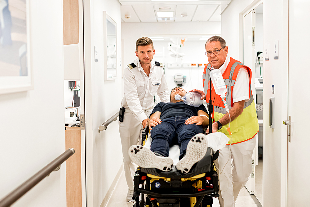 Flur des Bordhospitals. Ein männlicher Arzt und eine männliche Nurse schieben eine Liege mit einem Patienten drauf durch den Flur. 