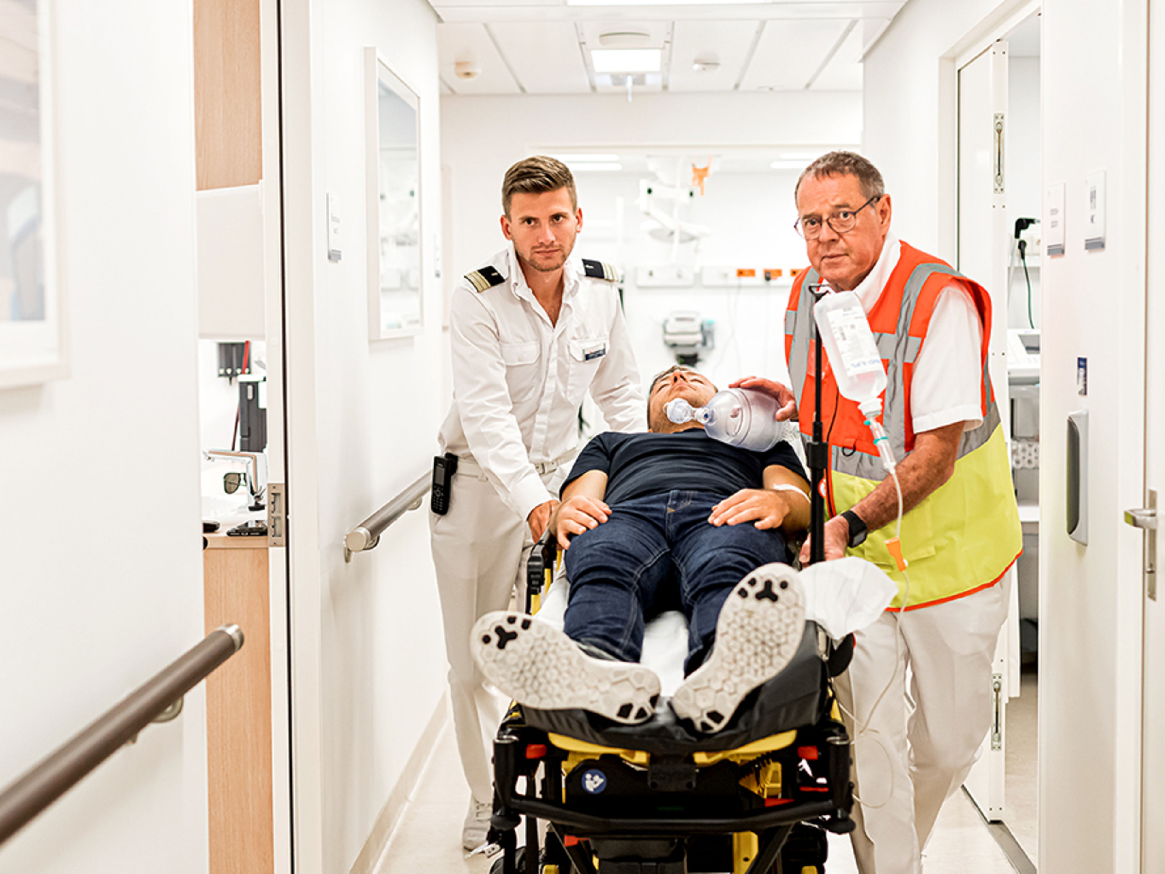 Flur des Bordhospitals. Ein männlicher Arzt und eine männliche Nurse schieben eine Liege mit einem Patienten drauf durch den Flur.