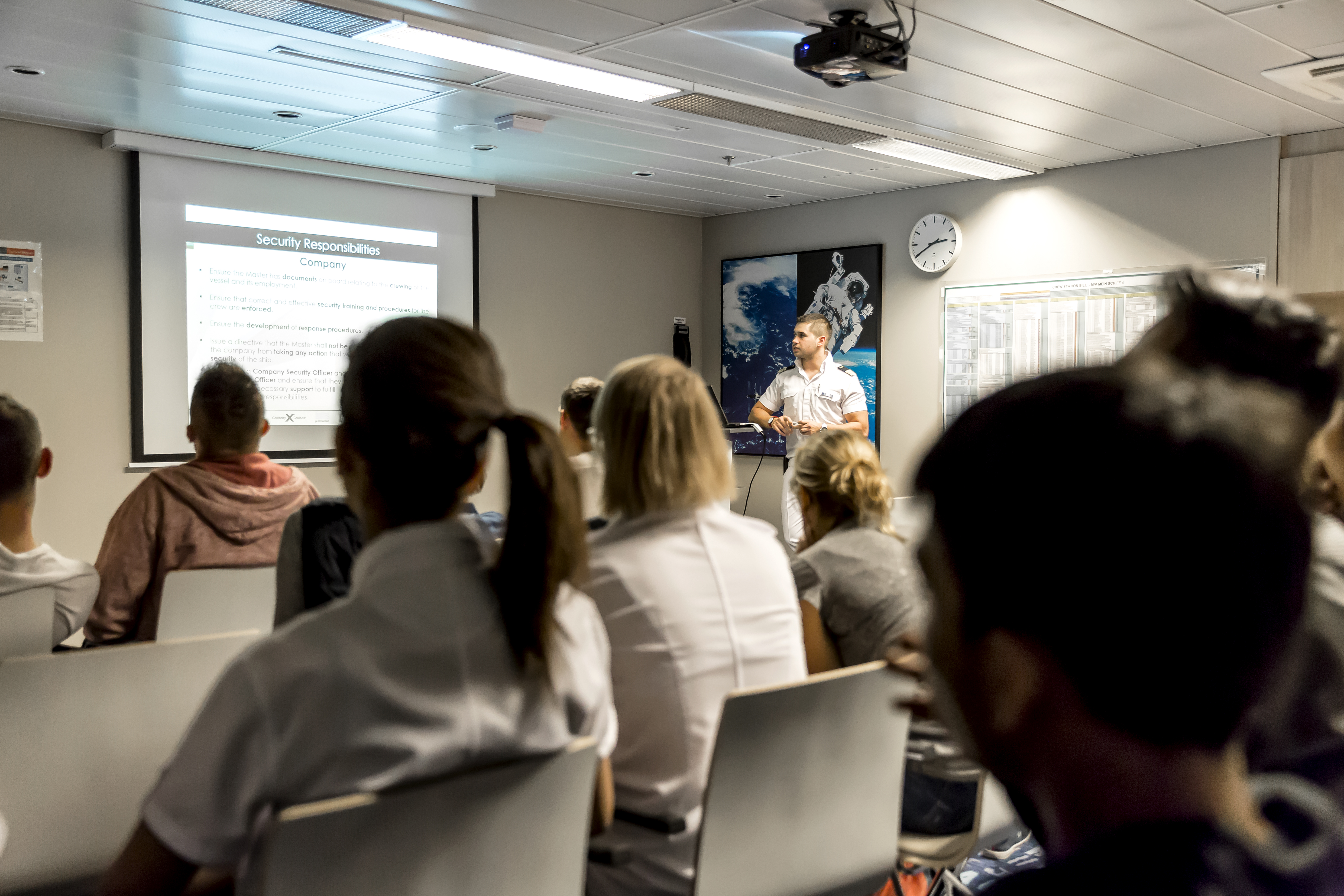 Crew-Mitglieder, die vor einem Beamer-Bild sitzen und einem Offizier zuhören 