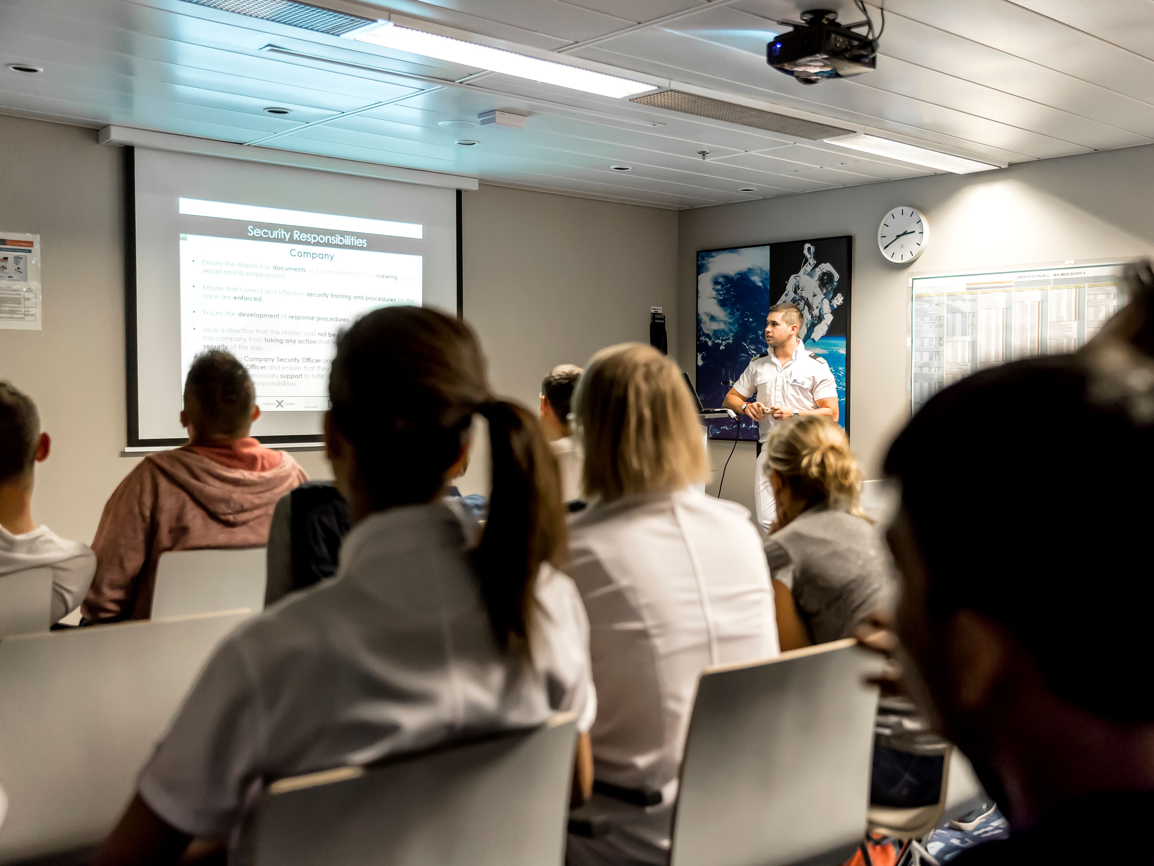 Crew-Mitglieder, die vor einem Beamer-Bild sitzen und einem Offizier zuhören