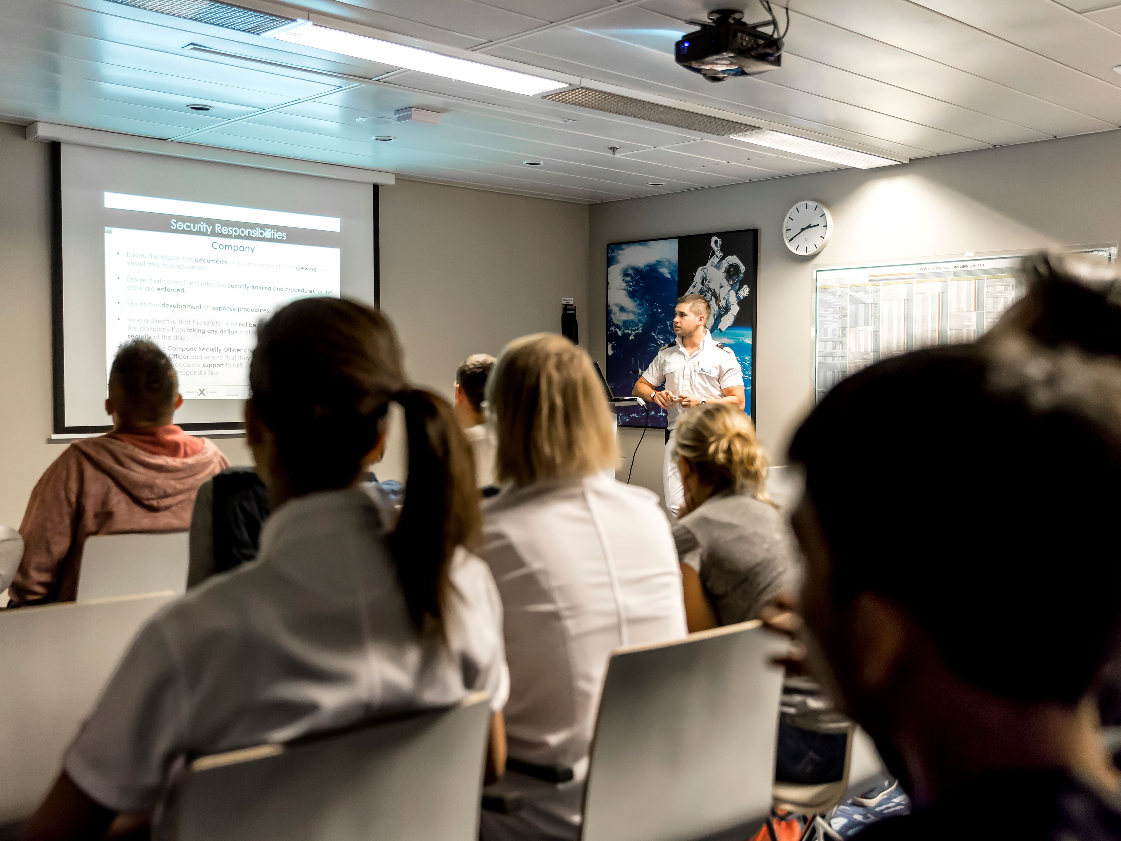Crew-Mitglieder, die vor einem Beamer-Bild sitzen und einem Offizier zuhören