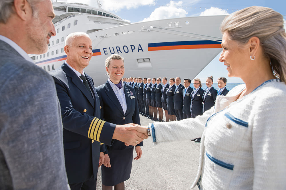 Spalier stehen vor der MS EUROPA. Davor gibt ein weiblicher Gast einem männlichen Offizier die Hand. 