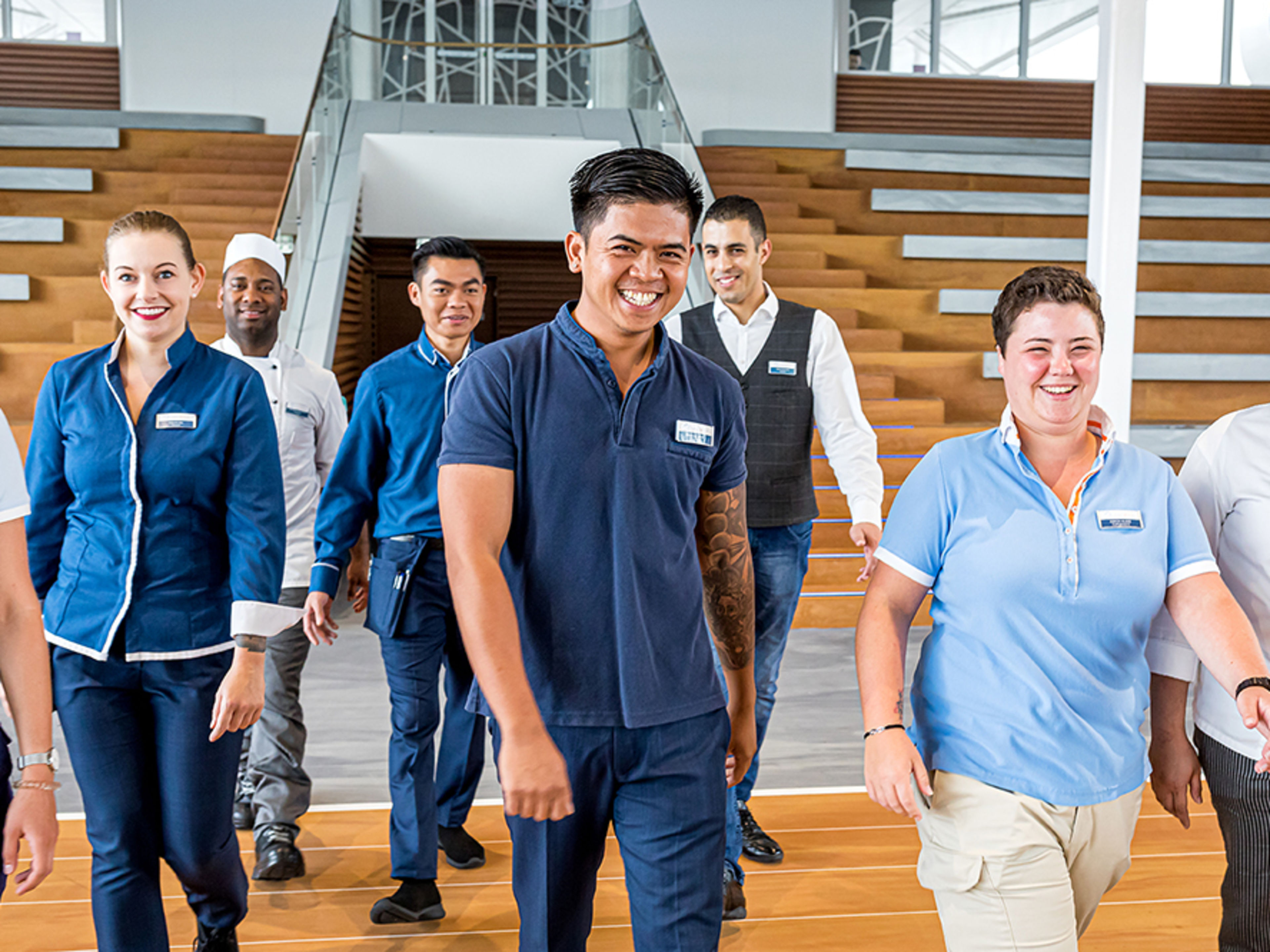 Arena. Acht verschiedene Crew Member in unterschiedlichen Uniformen und unterschiedlicher Nationalitäten gehen lächelnd auf die Kamera zu.