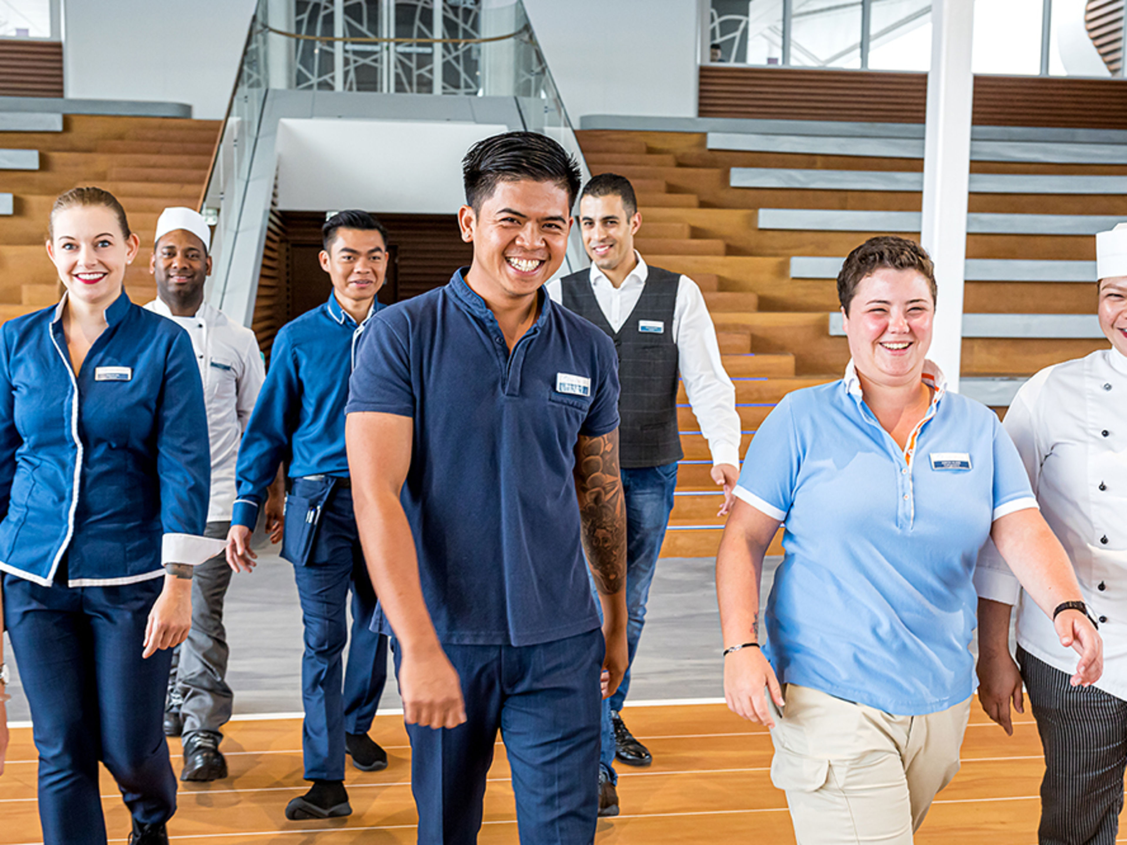Arena. Acht verschiedene Crew Member in unterschiedlichen Uniformen und unterschiedlicher Nationalitäten gehen lächelnd auf die Kamera zu.