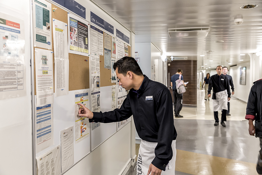Beschäftigter Flur im Crew Bereich. Ein männliches Crew Member steht an einem Aushang und überprüft die Trainingszeiten. 