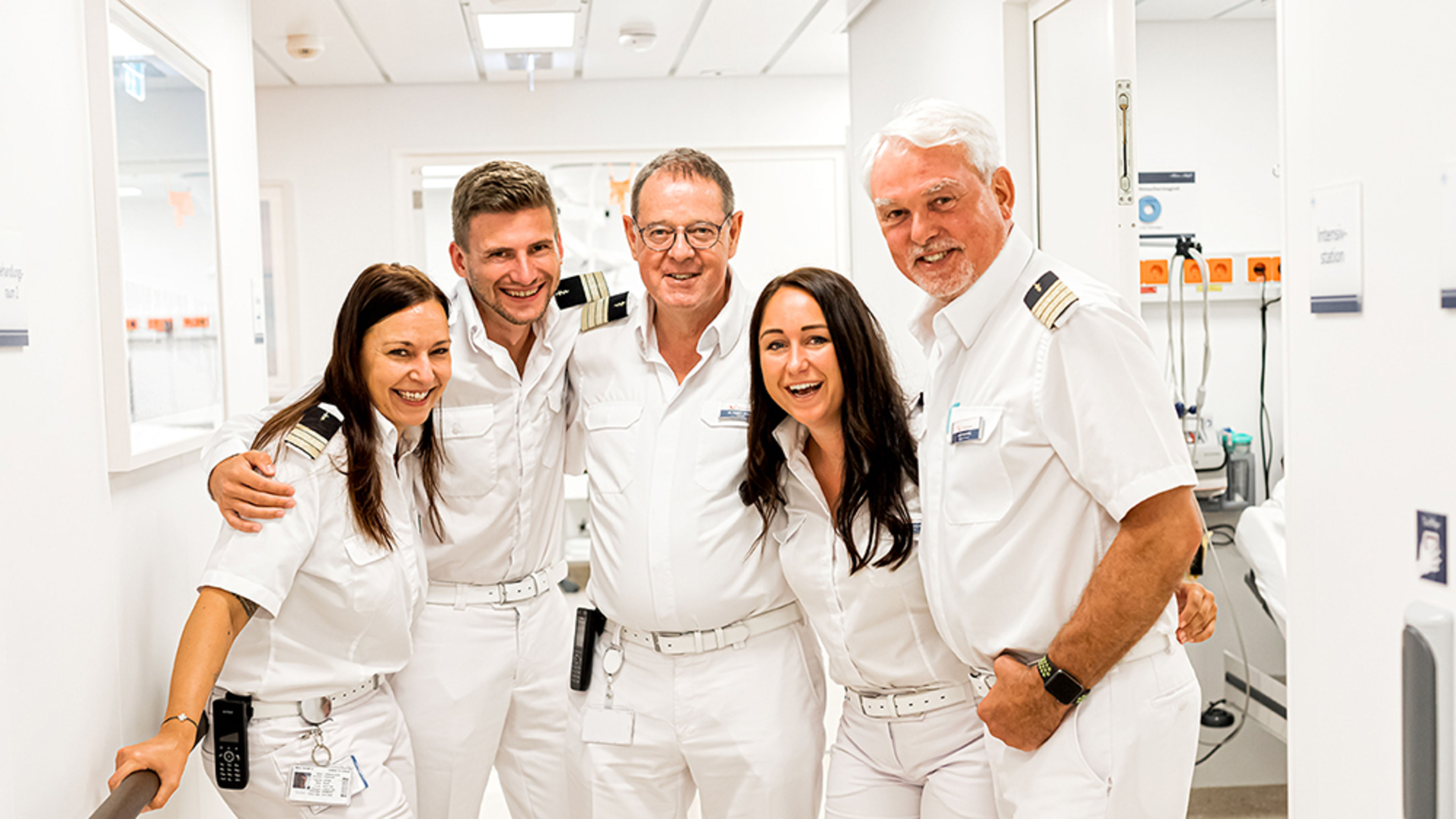 Flur im Bordhospital. Das fünfköpfige Hospitalteam steht Arm in Arm und lächelt in die Kamera.
