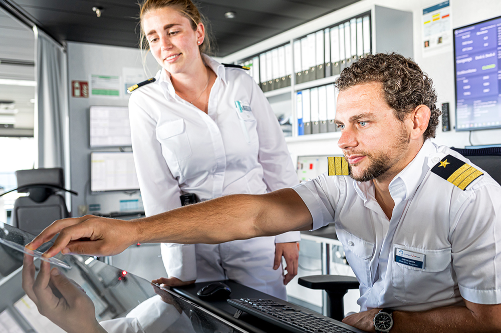 Schiffsbrücke. Zwei Offiziere sind im Bild. Ein männlicher Offizier sitzt an einem Schreibtisch und bedient ein Gerät. Eine weibliche Offizierin steht nehmen ihm und beobachtet, was er da macht. 