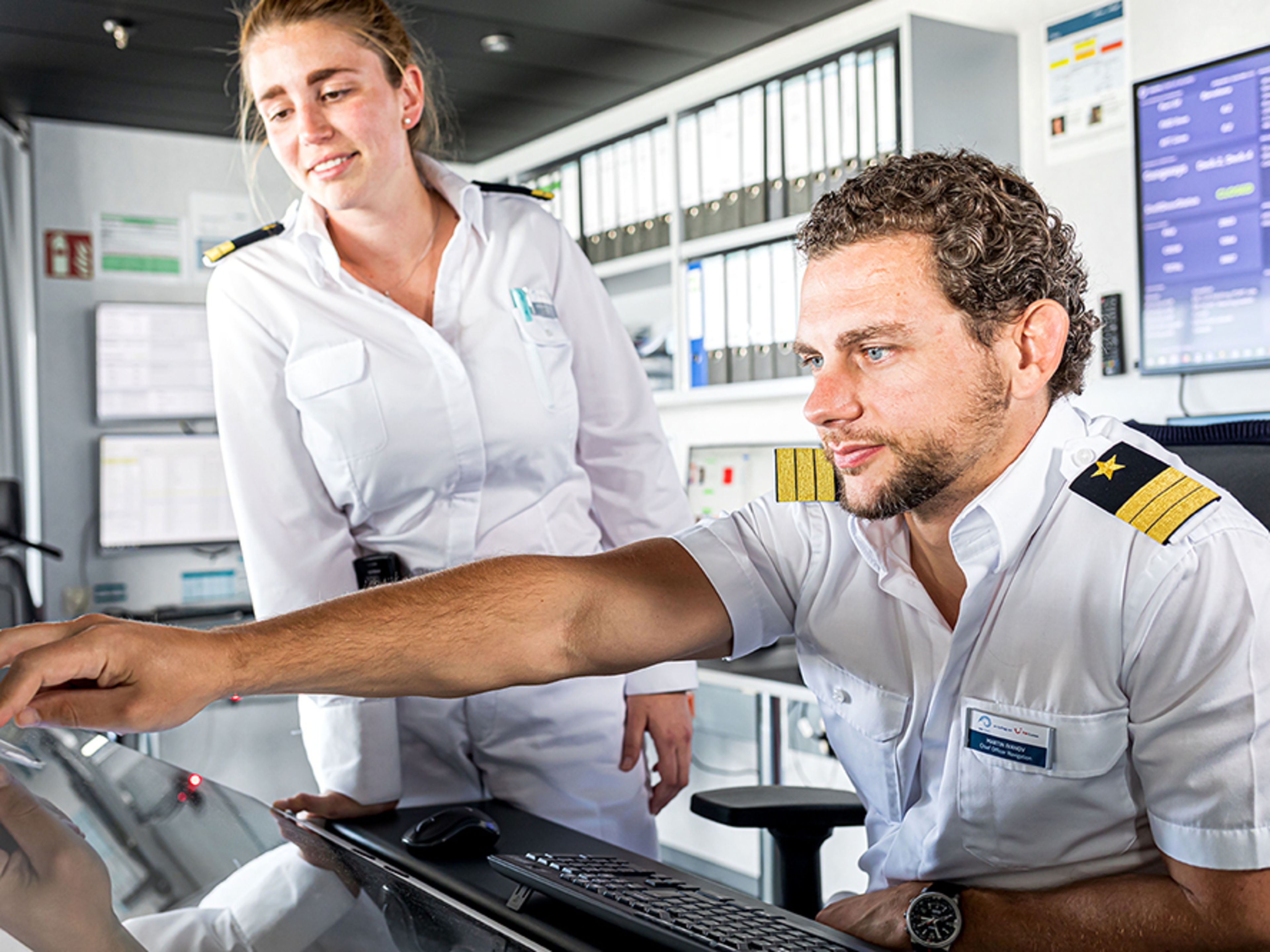 Schiffsbrücke. Zwei Offiziere sind im Bild. Ein männlicher Offizier sitzt an einem Schreibtisch und bedient ein Gerät. Eine weibliche Offizierin steht nehmen ihm und beobachtet, was er da macht.