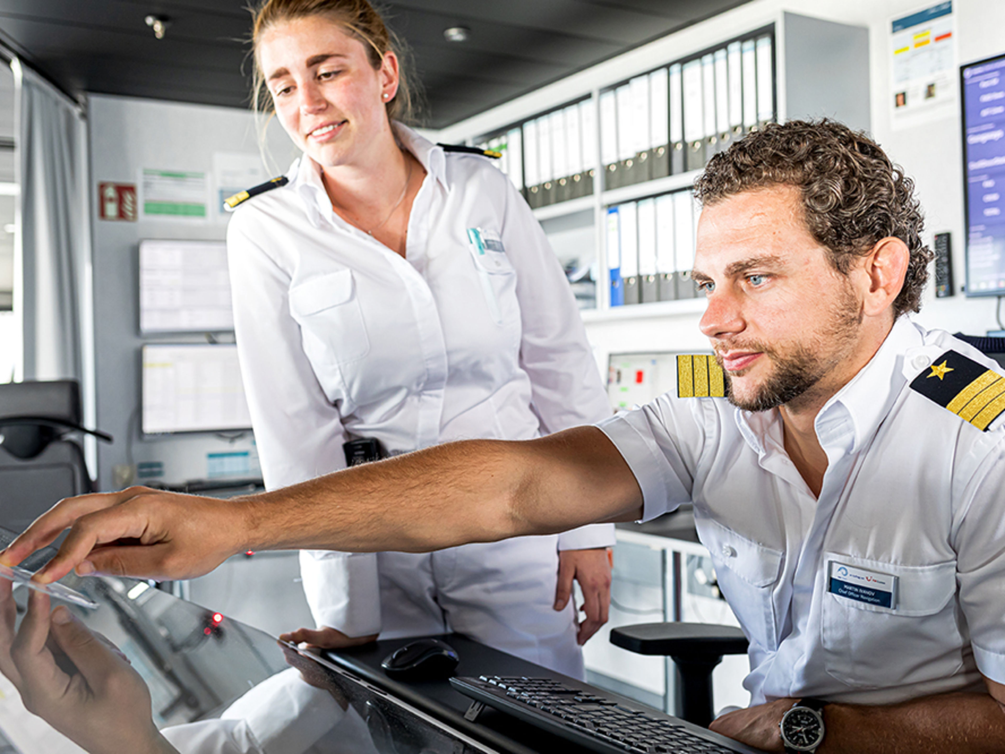 Schiffsbrücke. Zwei Offiziere sind im Bild. Ein männlicher Offizier sitzt an einem Schreibtisch und bedient ein Gerät. Eine weibliche Offizierin steht nehmen ihm und beobachtet, was er da macht.