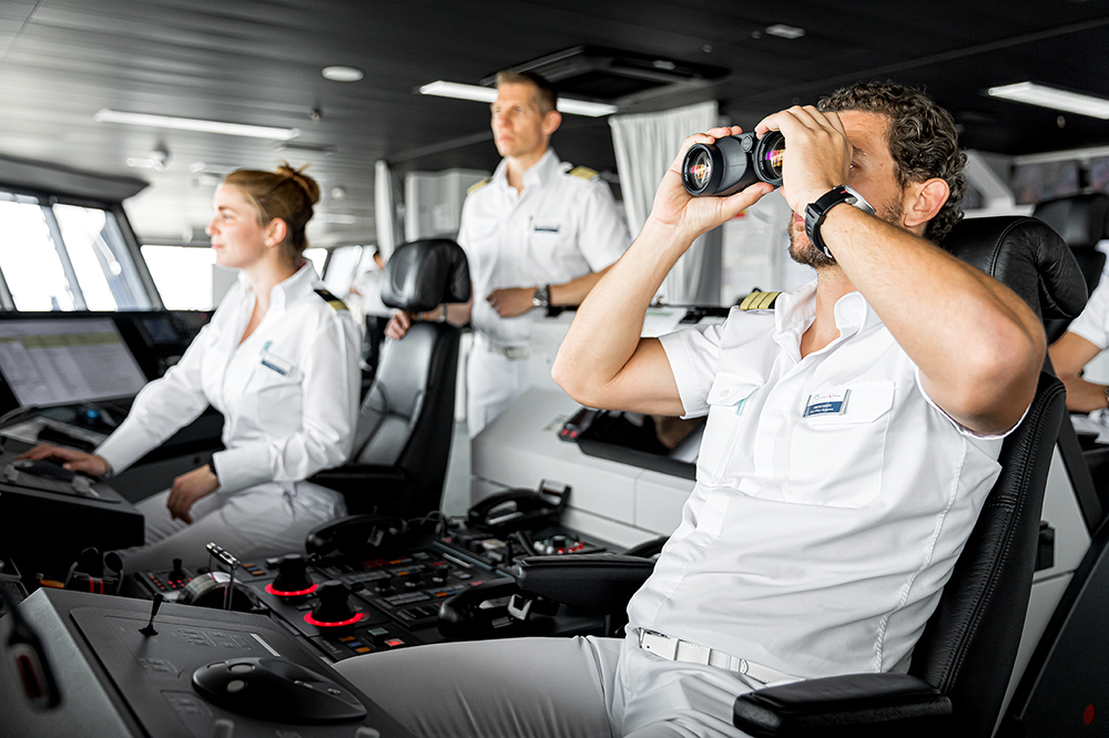 Auf der Schiffsbrücke. Drei Offiziere sind im Bild und halten Ausschau. Einer von ihnen schaut durch ein professionelles Fernglas. 