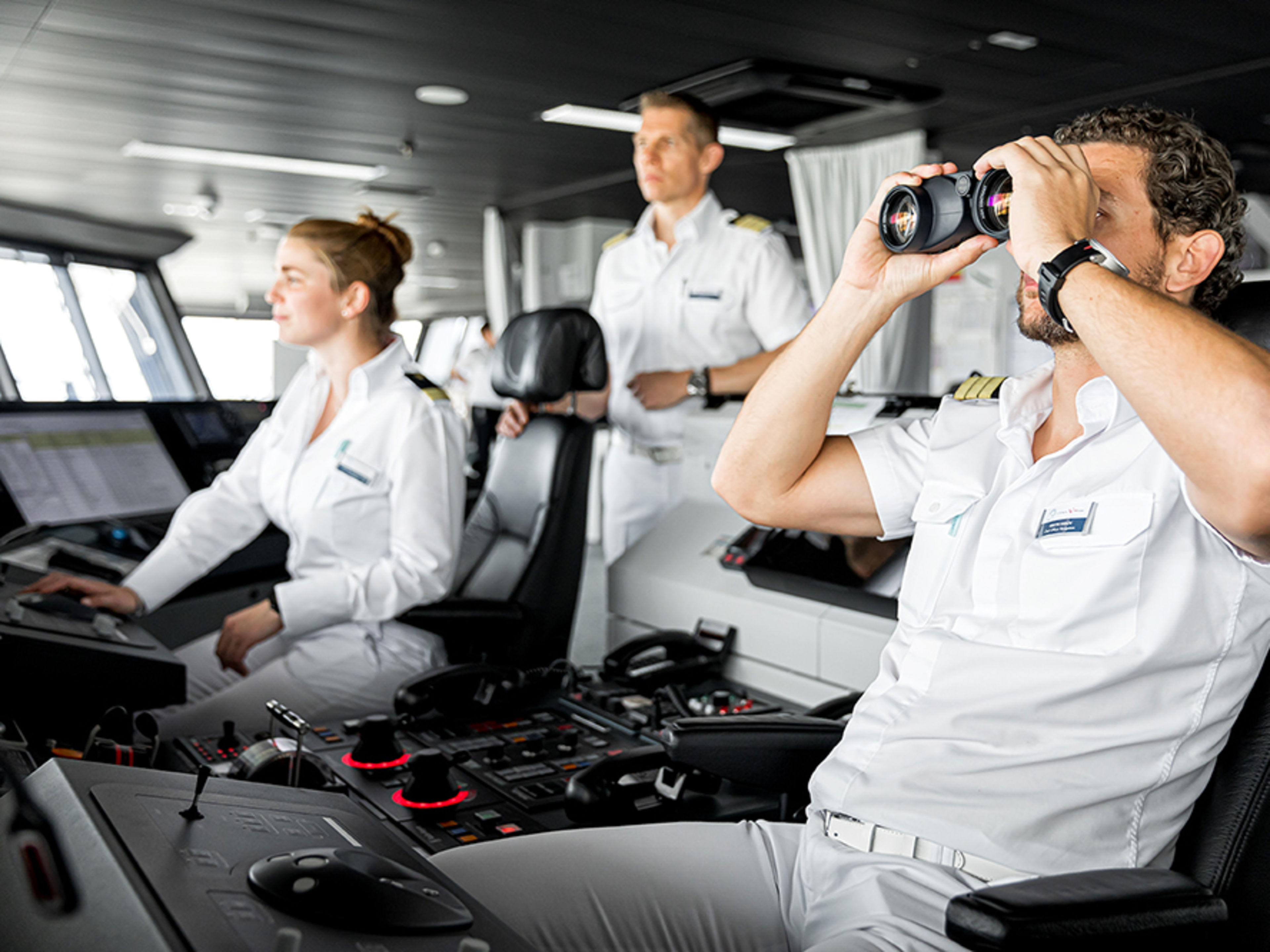 Auf der Schiffsbrücke. Drei Offiziere sind im Bild und halten Ausschau. Einer von ihnen schaut durch ein professionelles Fernglas.
