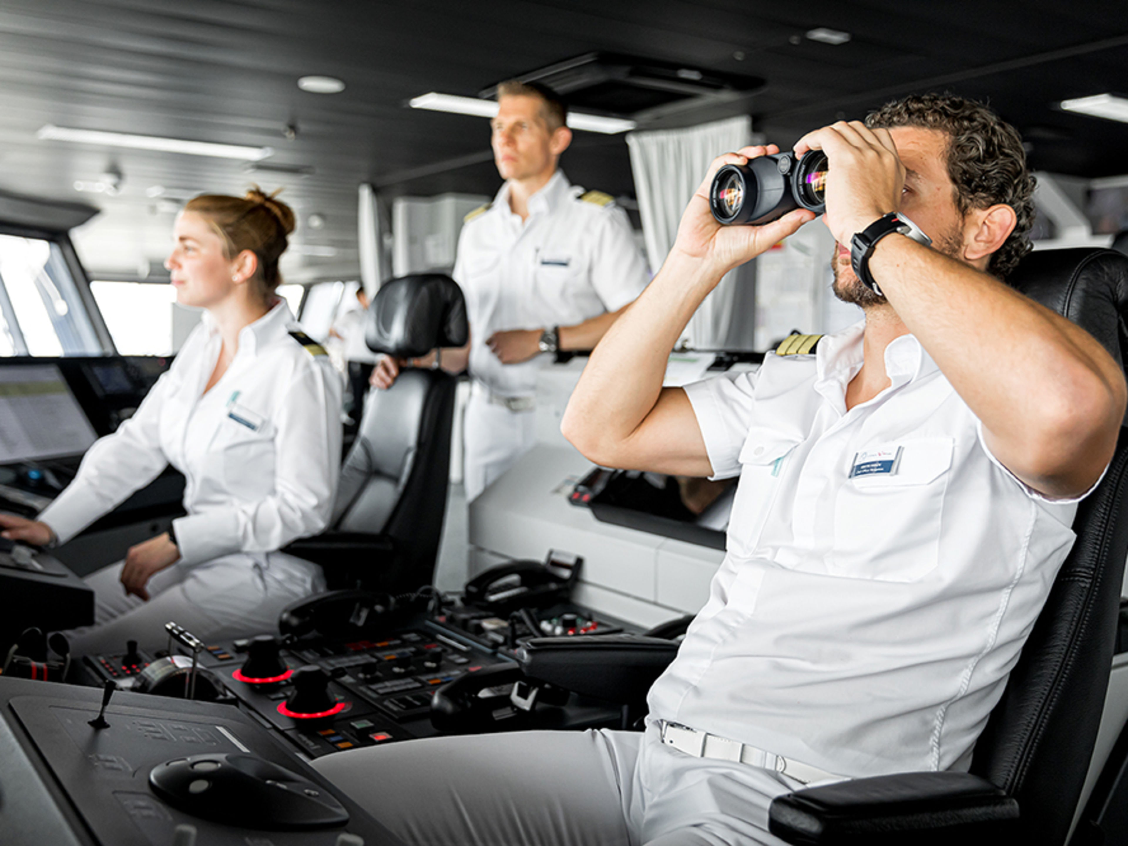 Auf der Schiffsbrücke. Drei Offiziere sind im Bild und halten Ausschau. Einer von ihnen schaut durch ein professionelles Fernglas.